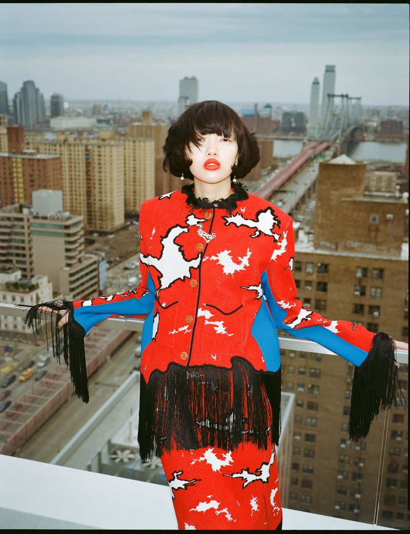 Model in a red and white patterned jacket with fringe standing on a rooftop overlooking a city skyline