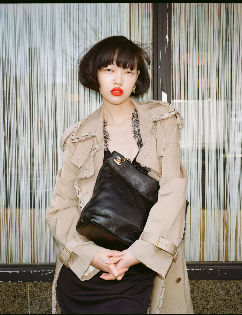 Model with dark bob haircut and red lipstick wearing a beige coat holding a black quilted handbag in front of vertical string curtains.
