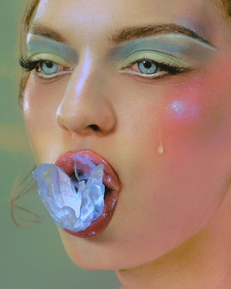 Close-up portrait of a woman with pastel blue eyeshadow and glossy lips holding a translucent resin sculpture in her mouth.