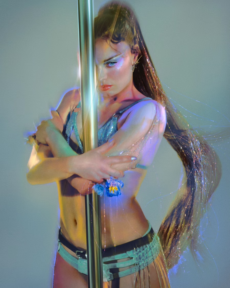 Woman with long dark hair and metallic bra posing beside a vertical pole against a neutral background