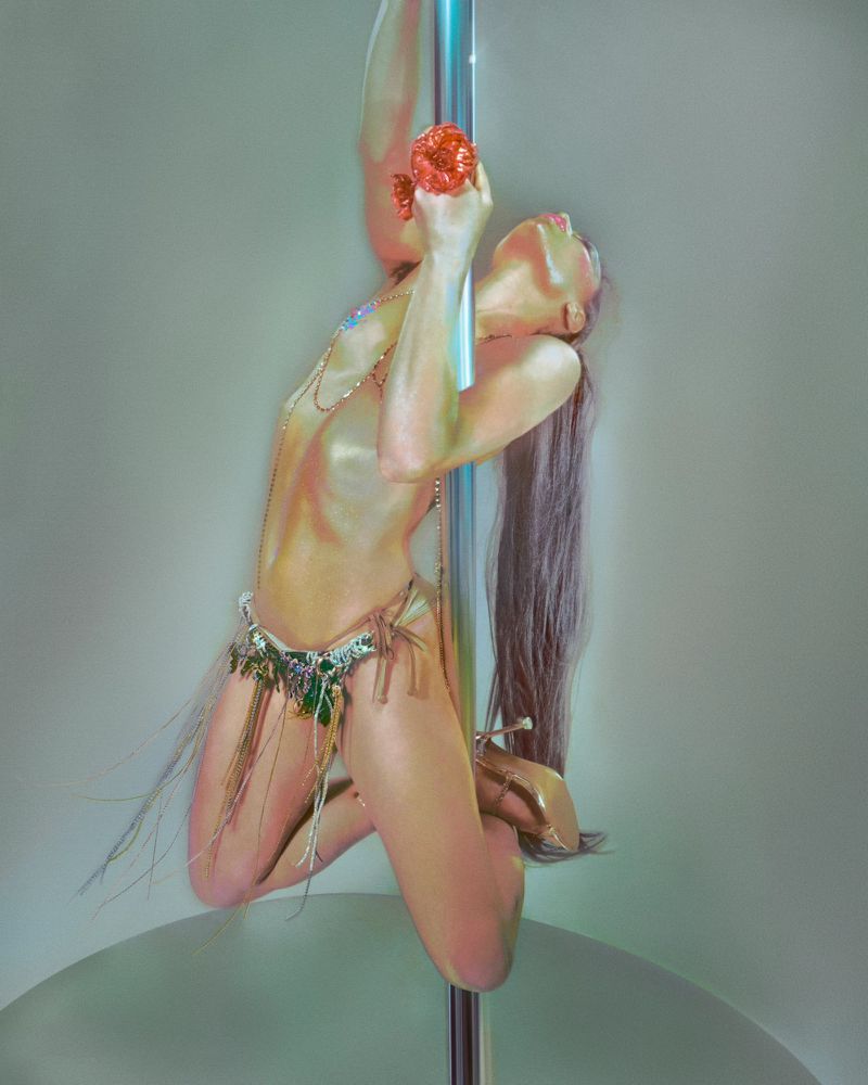 Model with long hair posing on a vertical chrome pole wearing embellished briefs and high heels against a pale background