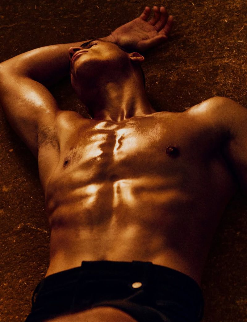 Shirtless male model lying down with arms raised under warm lighting for Numero Netherlands