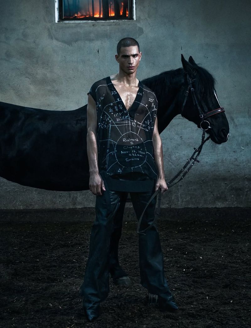 Man standing next to a black horse in a stable wearing a sheer printed top and dark trousers for Numero Netherlands.