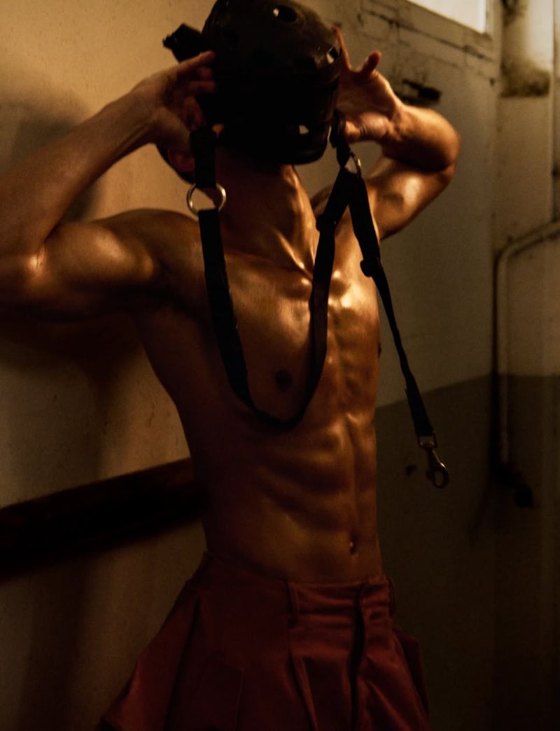 Shirtless male model wearing dark straps and red trousers holding a mask against a textured wall
