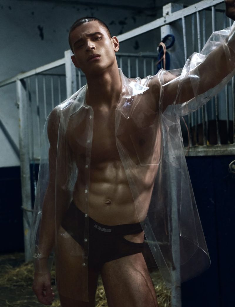 Male model in a transparent plastic shirt and dark briefs posing in an interior stall setting for Wild Narcissus.