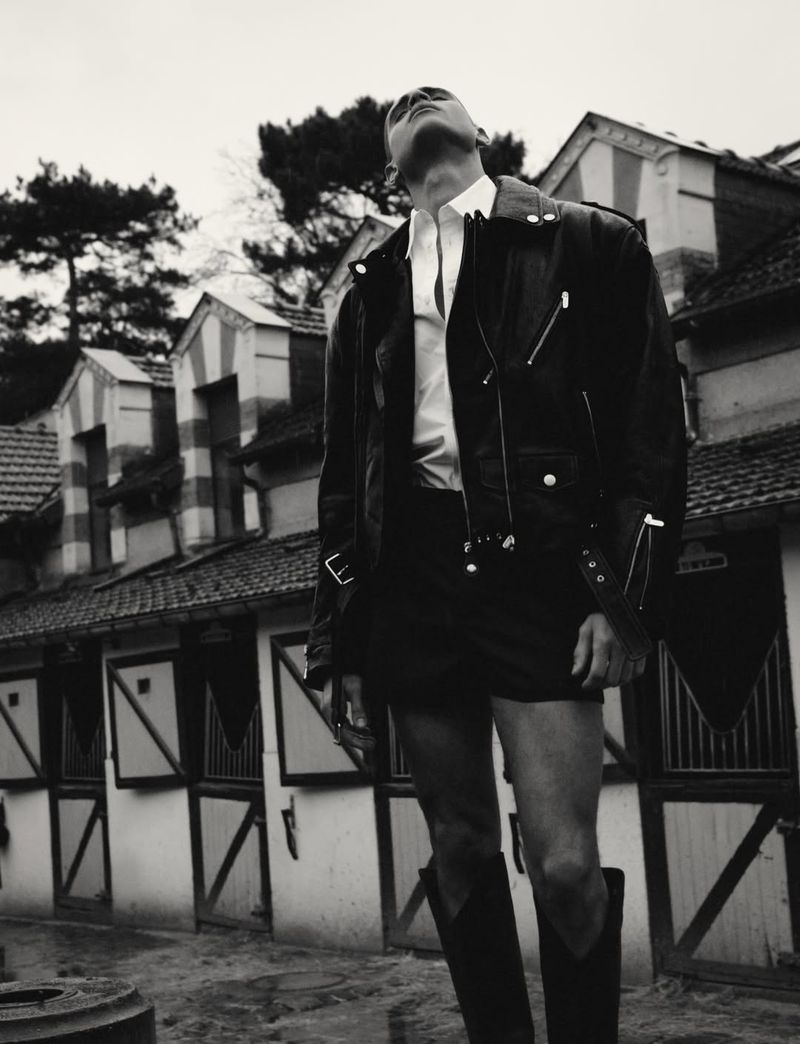 Model in black leather jacket and tall boots standing outdoors against a stable building exterior