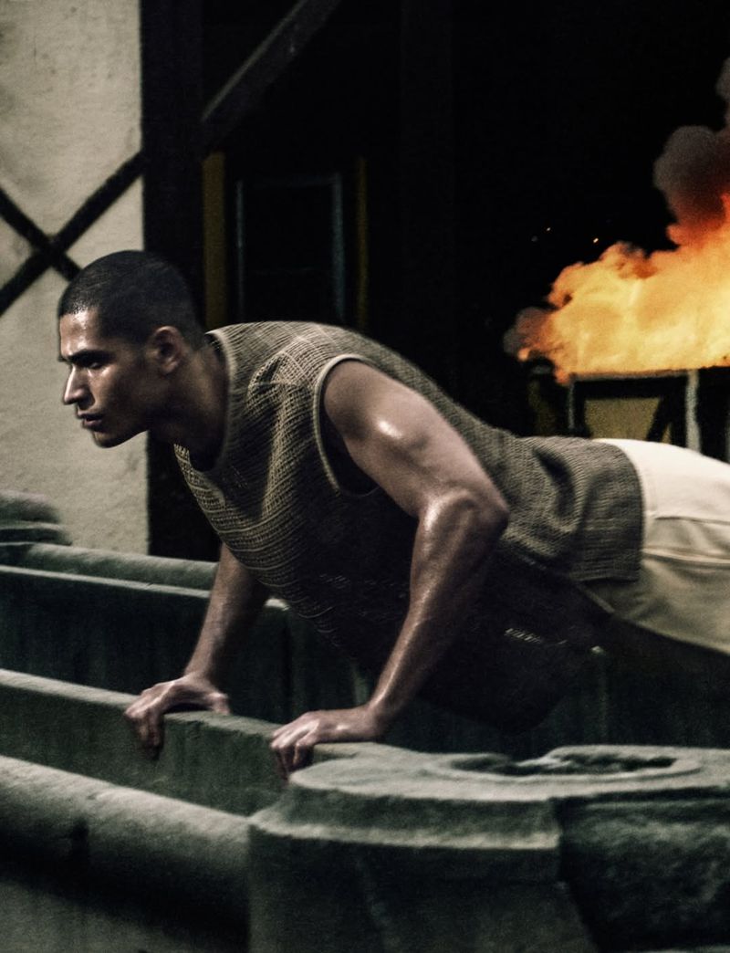 A man wearing a knit vest and shorts doing push-ups on stone structures with fire visible in the background.