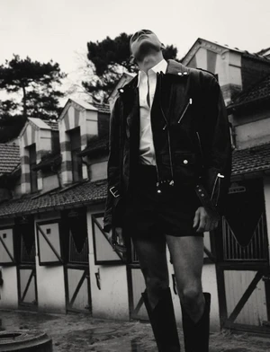 Model in black leather jacket and tall boots standing outdoors against a stable building exterior