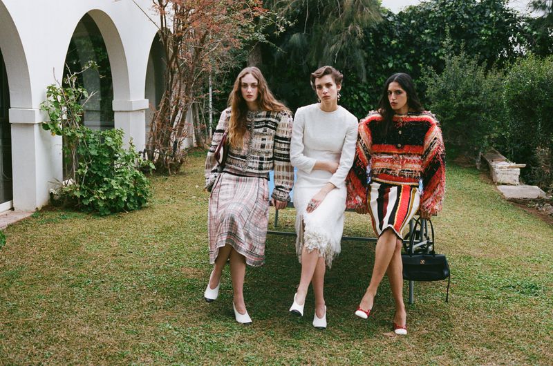Three female models posing seated together in a lush green garden wearing textured designer apparel for Marie Claire Greece.