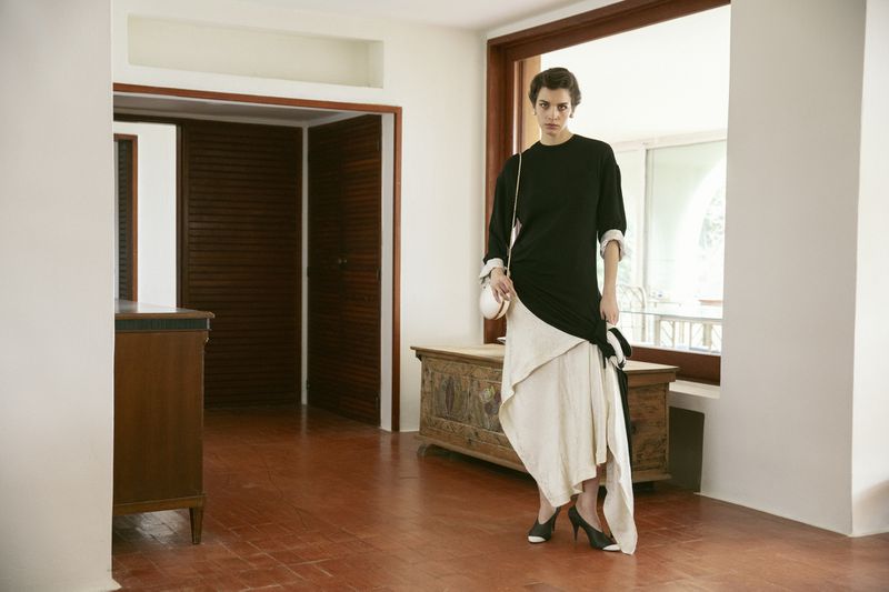 Model standing in an elegant interior wearing a black top and white skirt holding a small handbag
