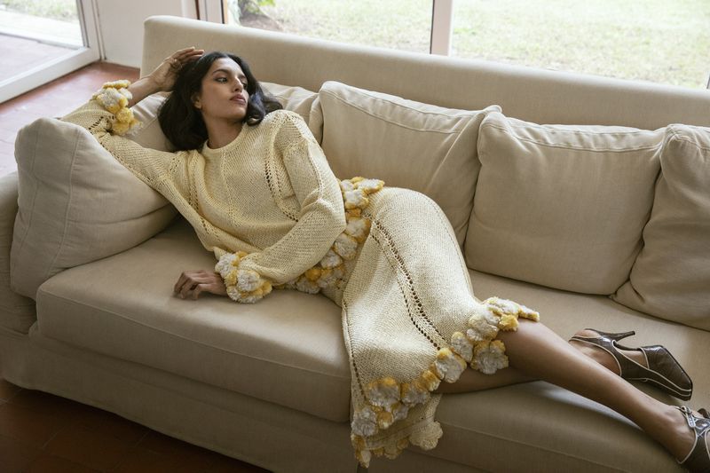 Model reclining on a beige sofa wearing a cream knit sweater and matching skirt with ruffled trim details