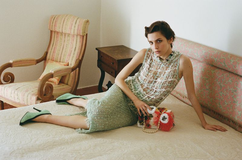 Model in green tweed skirt suit resting on a bed with a patterned armchair in a bedroom setting for Marie Claire Greece.