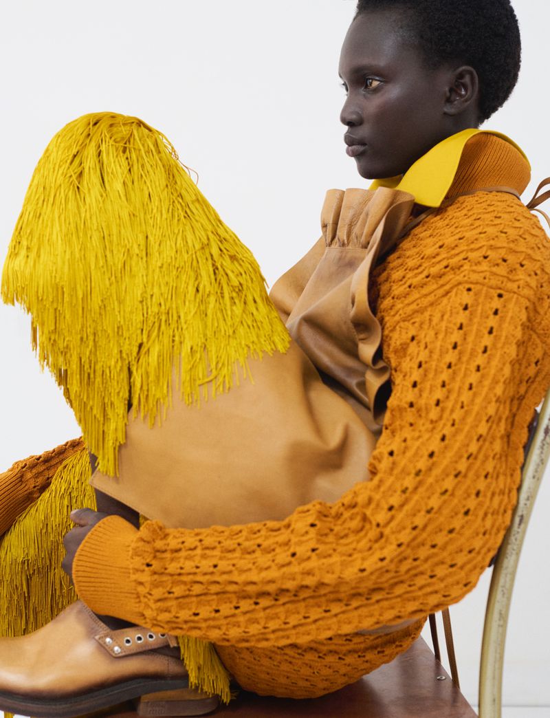 Aluel Keror in an orange textured sweater and yellow fringed skirt seated on a chair against a white background