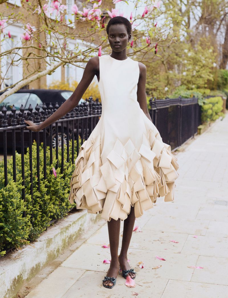 Model Aluel Keror in a sculptural cream dress standing on a sidewalk by a black fence for Exit Magazine.
