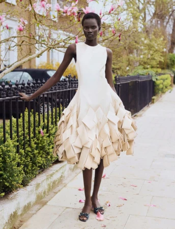 Model Aluel Keror in a sculptural cream dress standing on a sidewalk by a black fence for Exit Magazine.