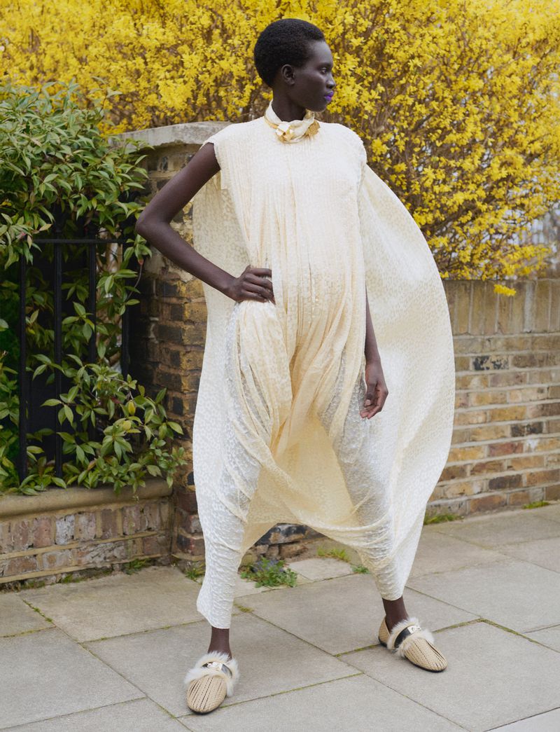 Model Aluel Keror wearing a cream textured tunic dress outdoors against a brick wall with yellow flowers for Exit Magazine
