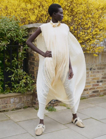 Model Aluel Keror wearing a cream textured tunic dress outdoors against a brick wall with yellow flowers for Exit Magazine