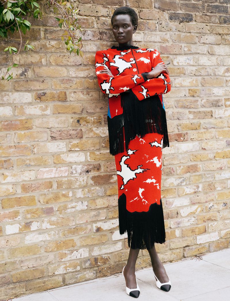 Model Aluel Keror in a red and black patterned fringe jacket and skirt leaning against a brick wall