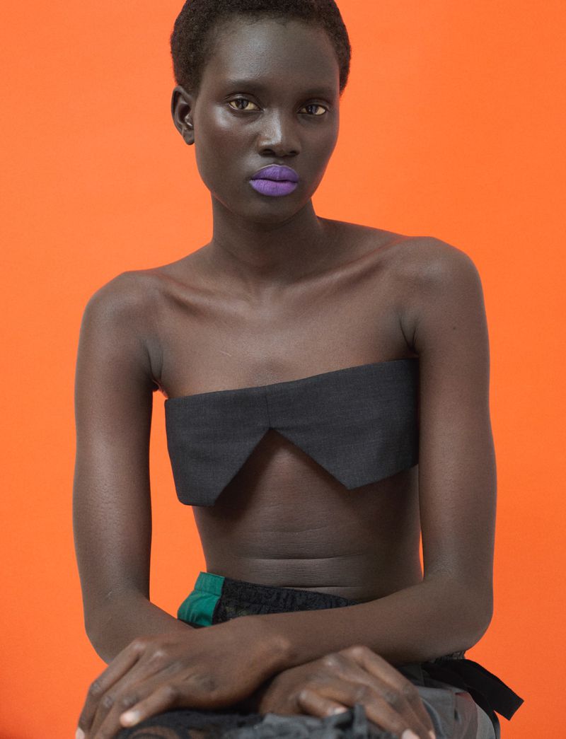 Model Aluel Keror wearing a black strapless top with vibrant purple lipstick against an orange backdrop for Exit Magazine