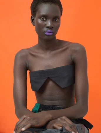Model Aluel Keror wearing a black strapless top with vibrant purple lipstick against an orange backdrop for Exit Magazine