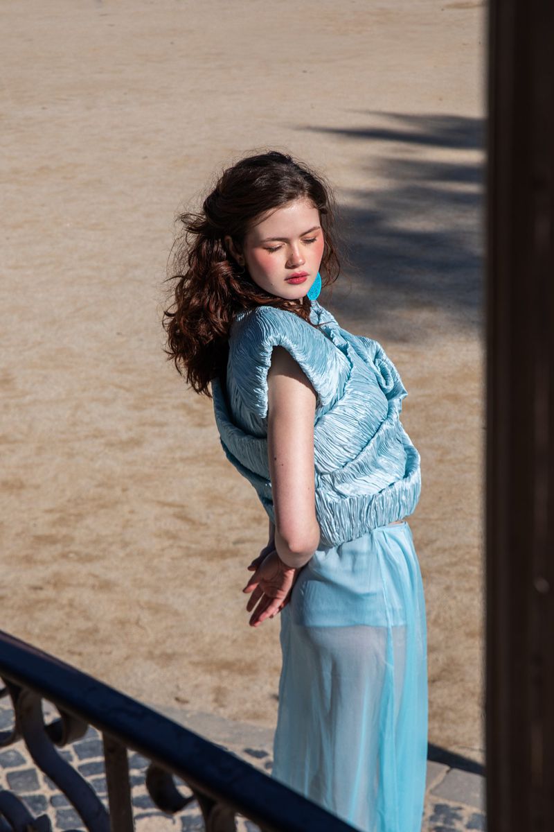 Model in a light blue textured top and sheer blue skirt standing outdoors in bright sunlight for Grazia Netherlands