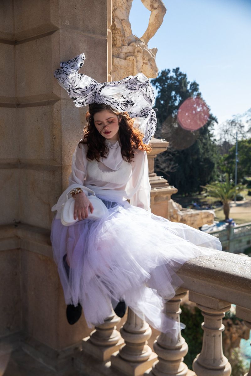 Woman in white sheer blouse and lavender tulle skirt wearing a large patterned headdress while sitting on a stone balustrade