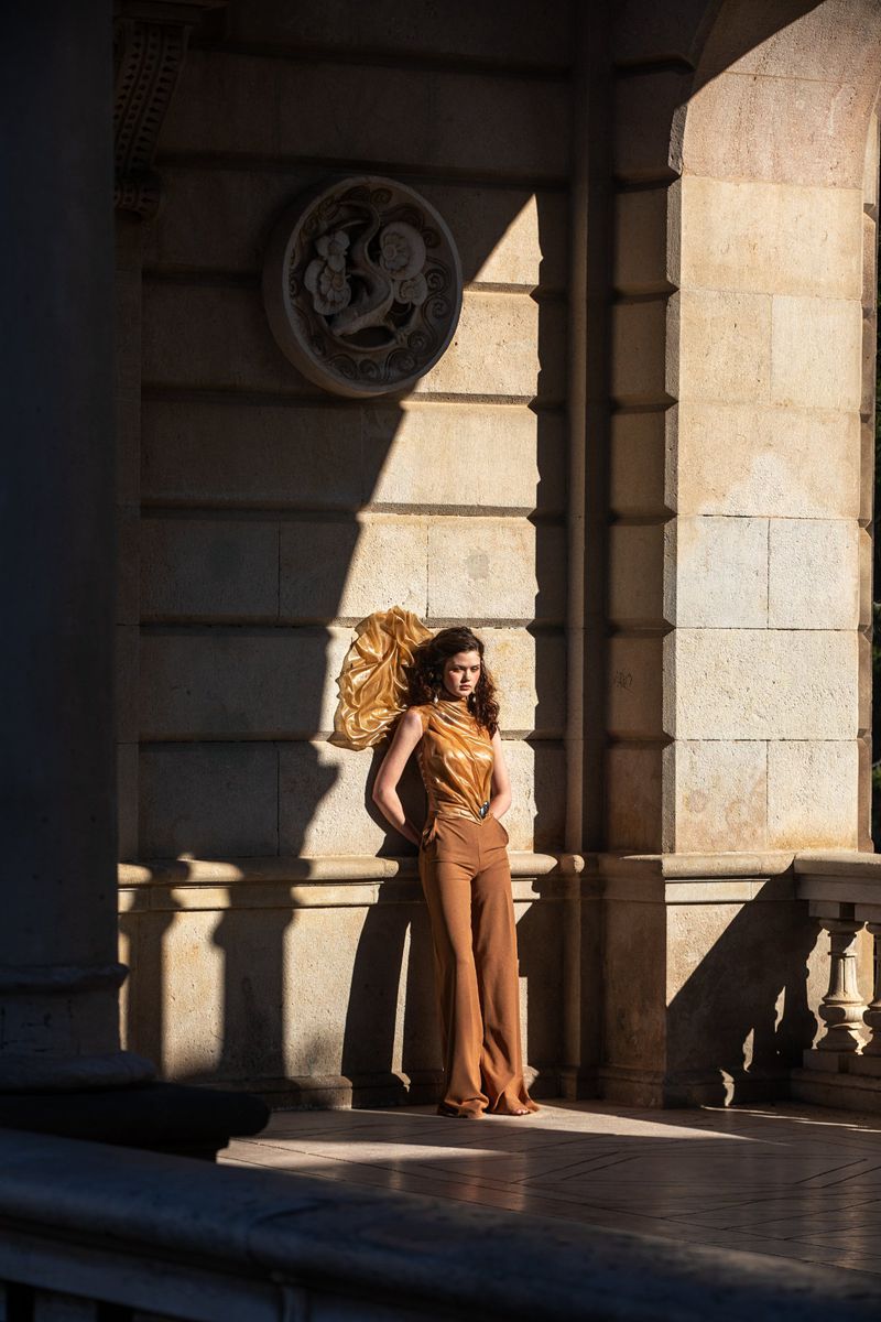 Woman in brown trousers and top leaning against a classical stone wall under strong sunlight, for Grazia Netherlands.