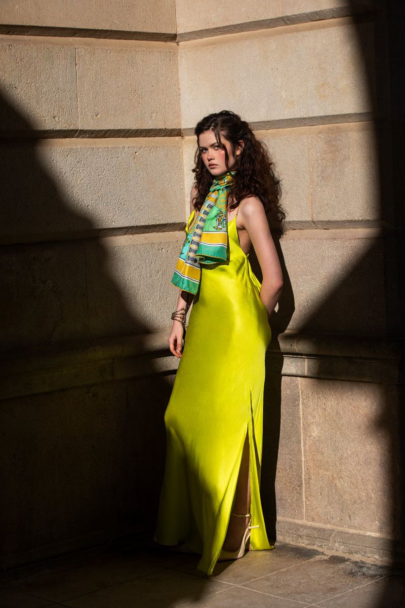 Woman in bright yellow slip dress wearing a patterned green scarf standing in an architectural corner under sunlight