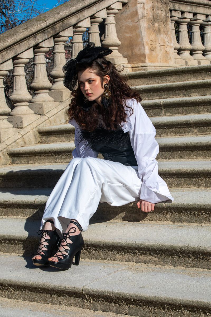 Model seated on stone stairs wearing a white shirt, black top, and lace-up heels with a dark fascinator.