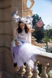 Woman in white sheer blouse and lavender tulle skirt wearing a large patterned headdress while sitting on a stone balustrade