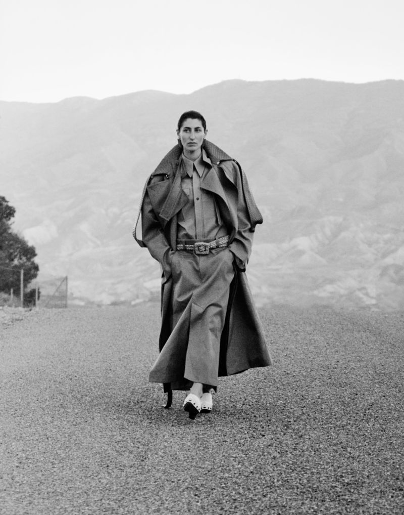 Model walking forward on an open road wearing a long belted trench coat, with hazy distant mountains in the background.