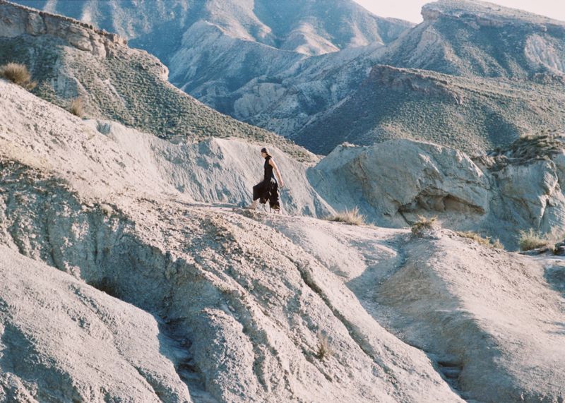 Model in black clothing walking along a rugged desert ridge surrounded by barren mountain terrain