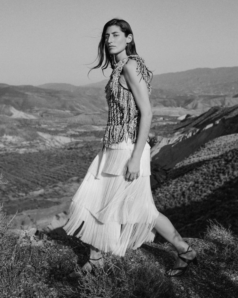 Black and white portrait of a female model in a textured fringed top and layered skirt standing on a rocky hillside landscape.