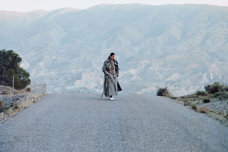 Model wearing a long grey coat walking on an asphalt road with mountains in the background