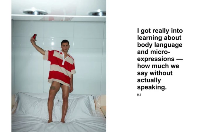 Man stands on a bed in a polo shirt holding a cell phone in a brightly lit room with white bedding and walls. Interior shot.
