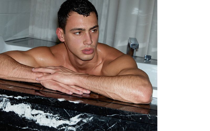 Close-up of an adult male resting in a bathtub, showcasing his physique and a glimpse of the bathroom interior.