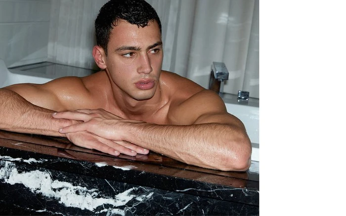Model Barak Shamir leaning on the edge of a black and white marble bathtub with arms crossed in a bathroom portrait.