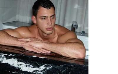 Model Barak Shamir leaning on the edge of a black and white marble bathtub with arms crossed in a bathroom portrait.