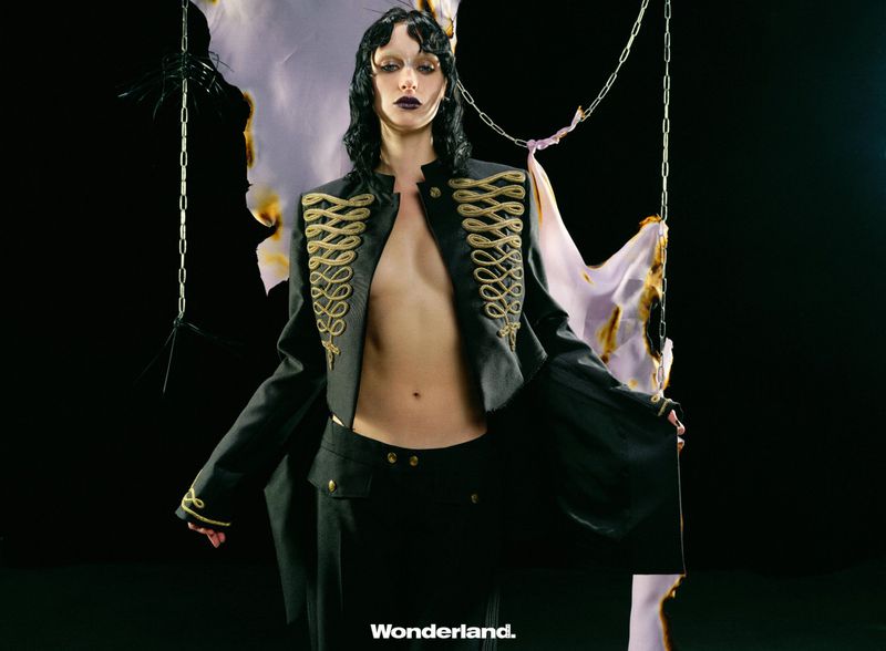 Model in black military-style jacket with gold embroidery posing against a dark background with hanging chains and burned fabric