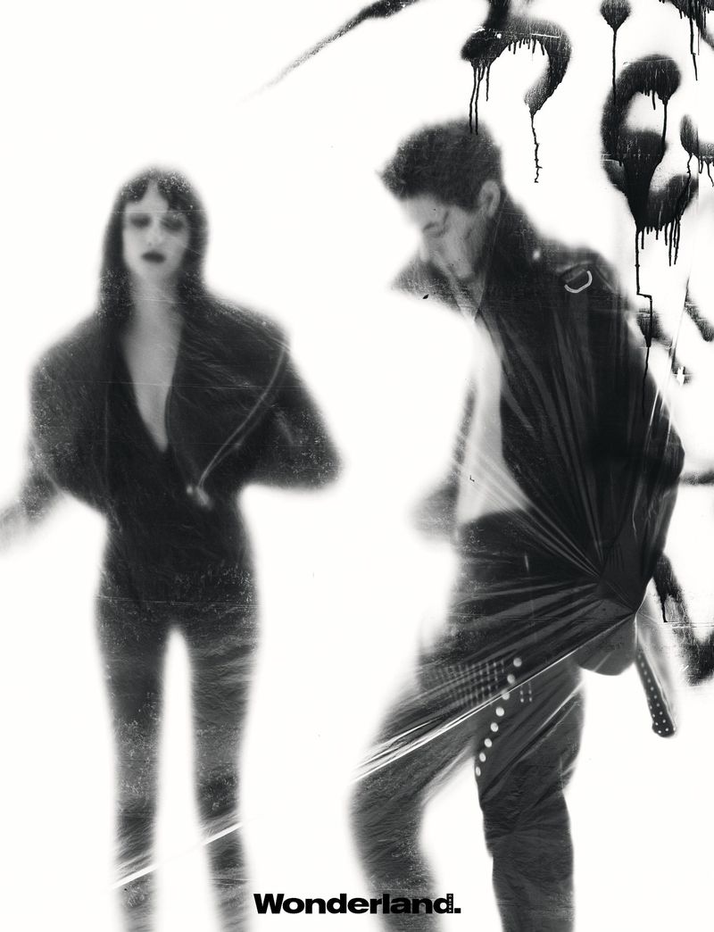 Black and white fashion editorial image featuring two models in dark jackets against a high-key backdrop with graffiti elements.