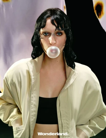 Model with dark hair and pale eyebrows blowing a bubblegum bubble while wearing a beige zip jacket for Wonderland China.