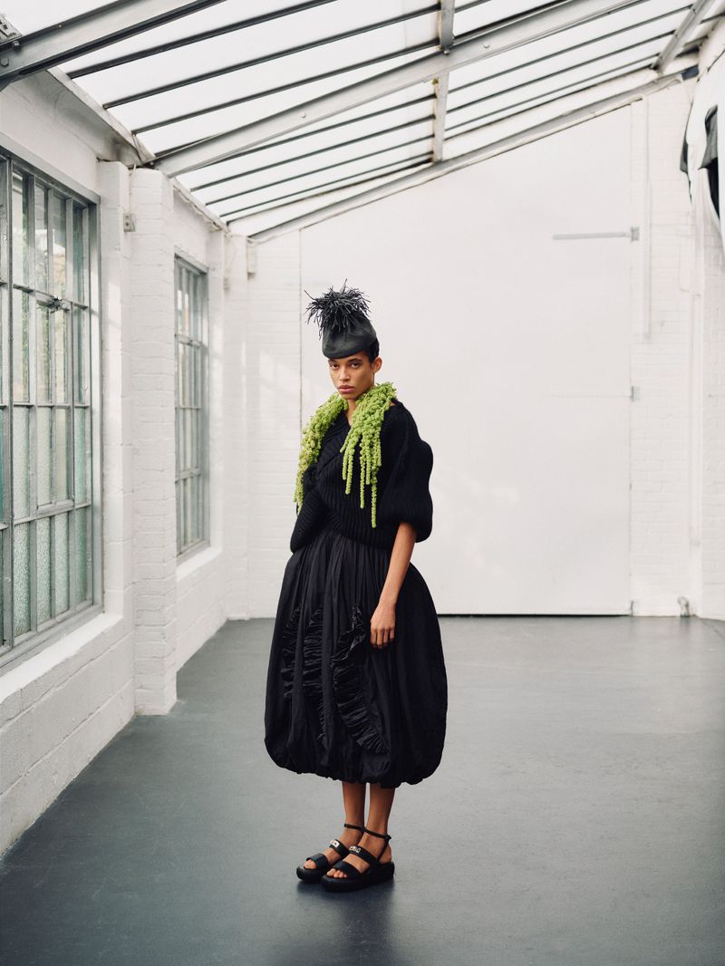 Model wearing a voluminous black dress and textured green scarf stands in a bright industrial studio space.