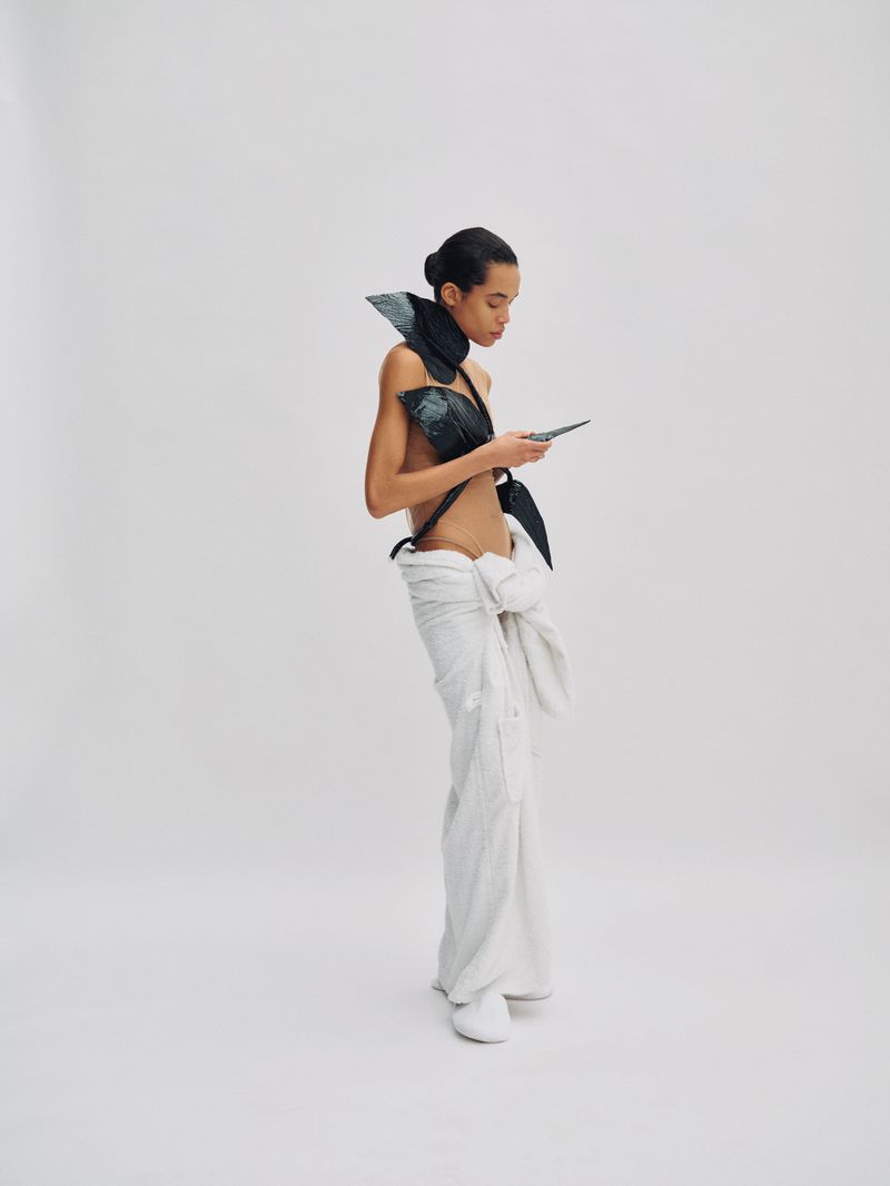 Model Lara Menezes wearing sculptural black top and white trousers against a white seamless studio backdrop.