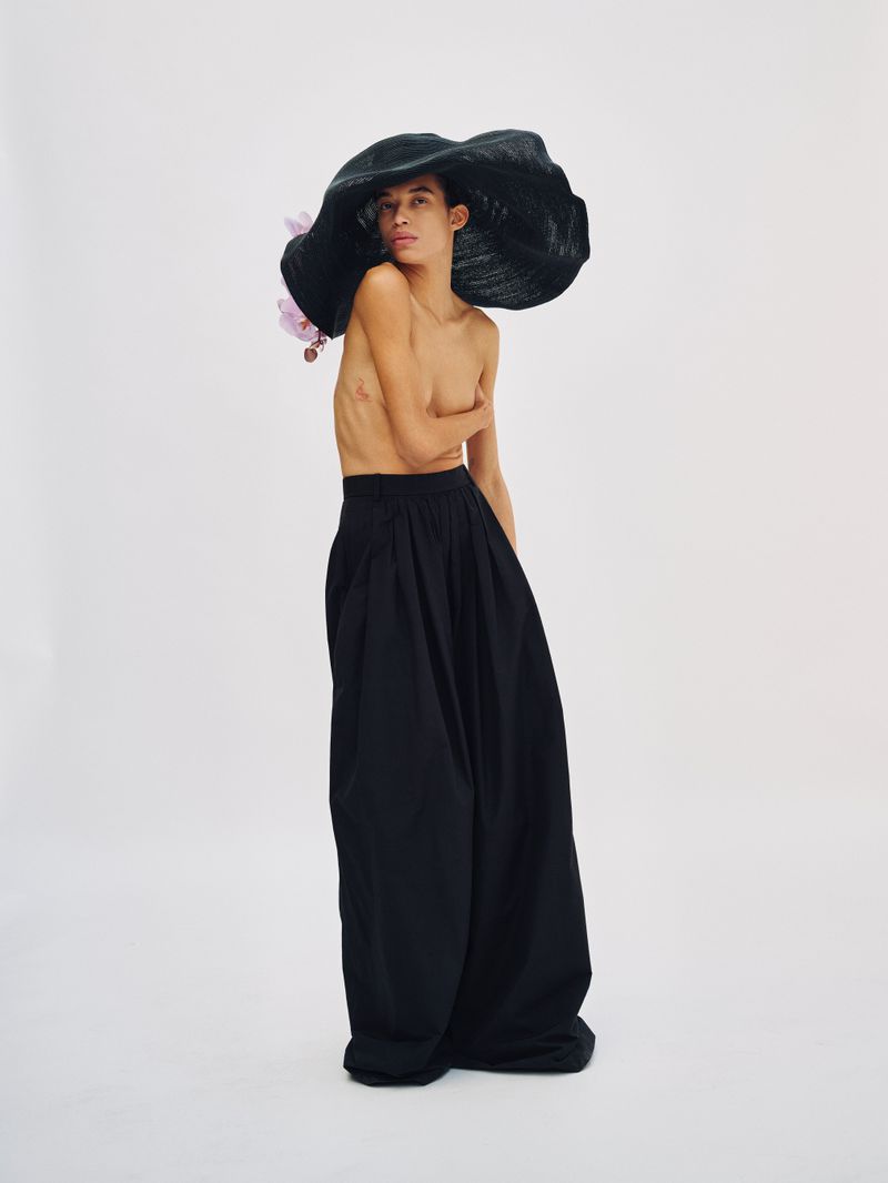Model wearing an oversized black hat with a pink flower and black trousers against a white backdrop