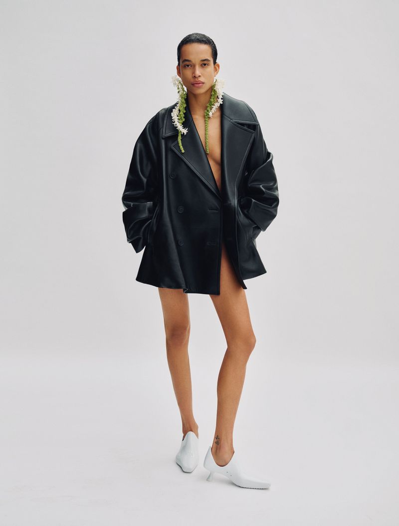 Model Lara Menezes wearing an oversized black leather jacket and white shoes against a plain white background for Exit Magazine.
