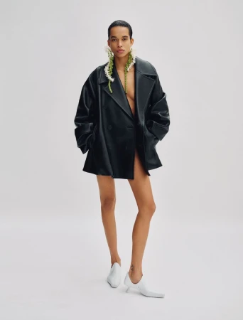 Model Lara Menezes wearing an oversized black leather jacket and white shoes against a plain white background for Exit Magazine.