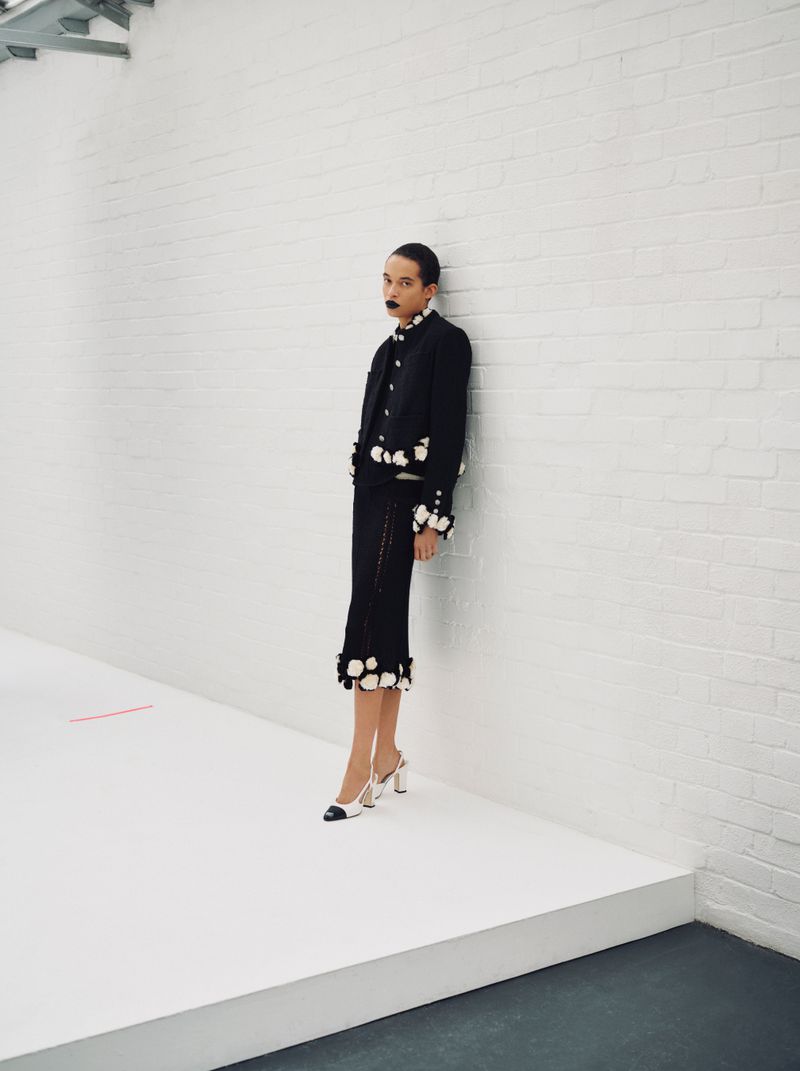Model leaning against a white brick wall wearing a black jacket and matching skirt with white floral accents.