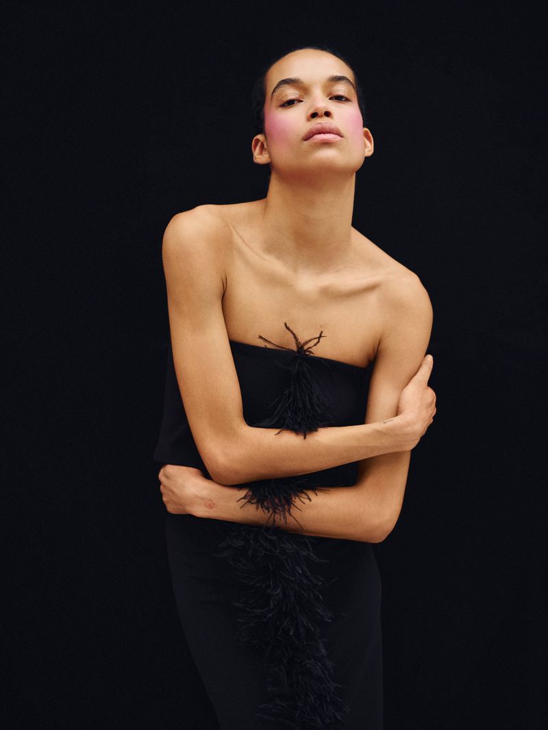 Model with pink blush poses in a black strapless dress with feather trim against a dark background, fashion photography in studio.