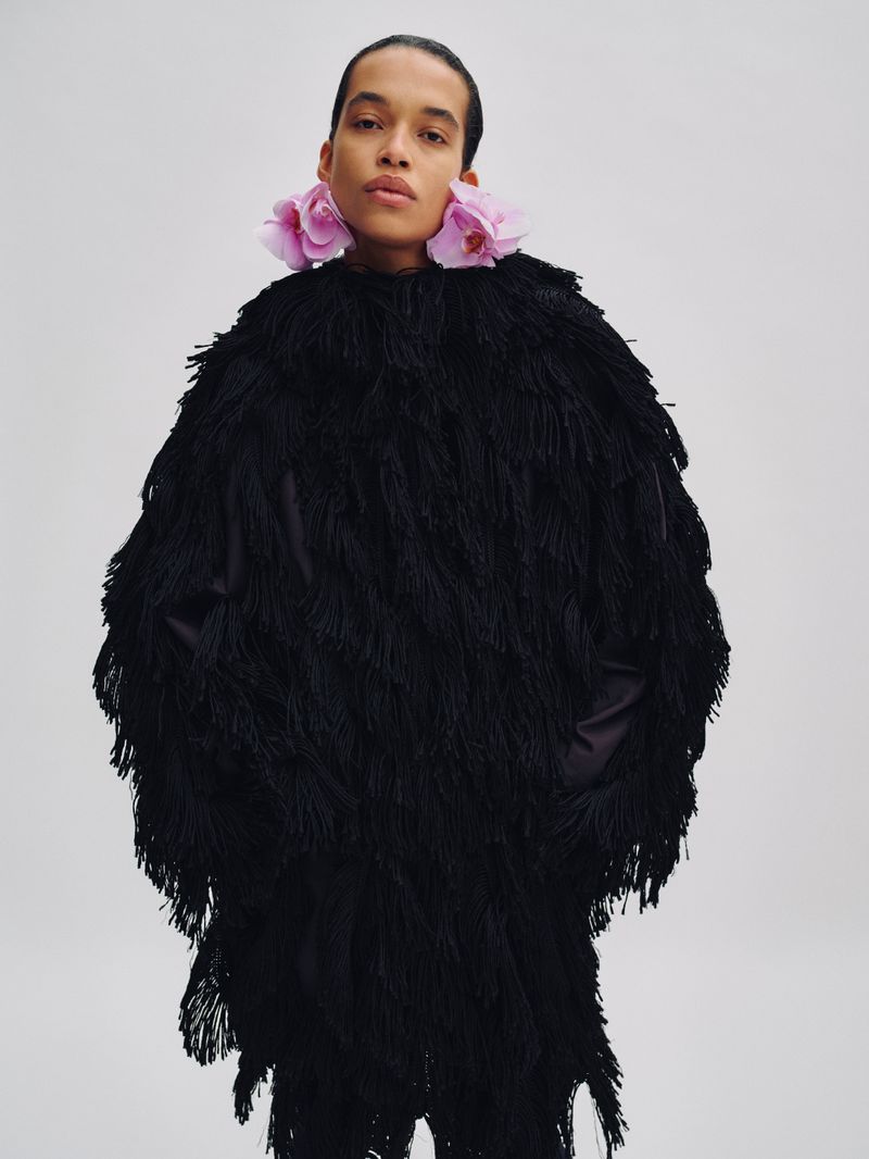 Model wearing a black fringed coat with large pink orchid flowers tucked behind ears against white background