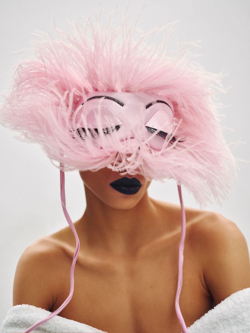 Model wearing a feathered pink mask with painted eyes and dark lipstick, photographed against a white background.